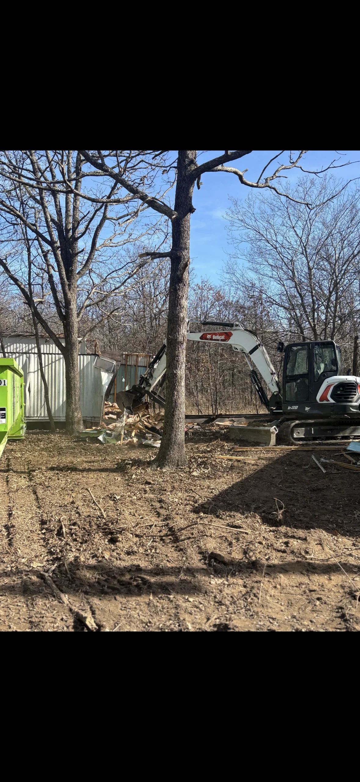 Bobcat excavator grading residential lot with mature trees and metal storage shed